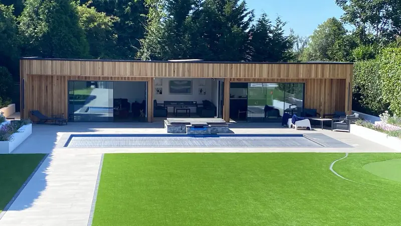Timber-clad garden building with wide glazed openings beside a swimming pool